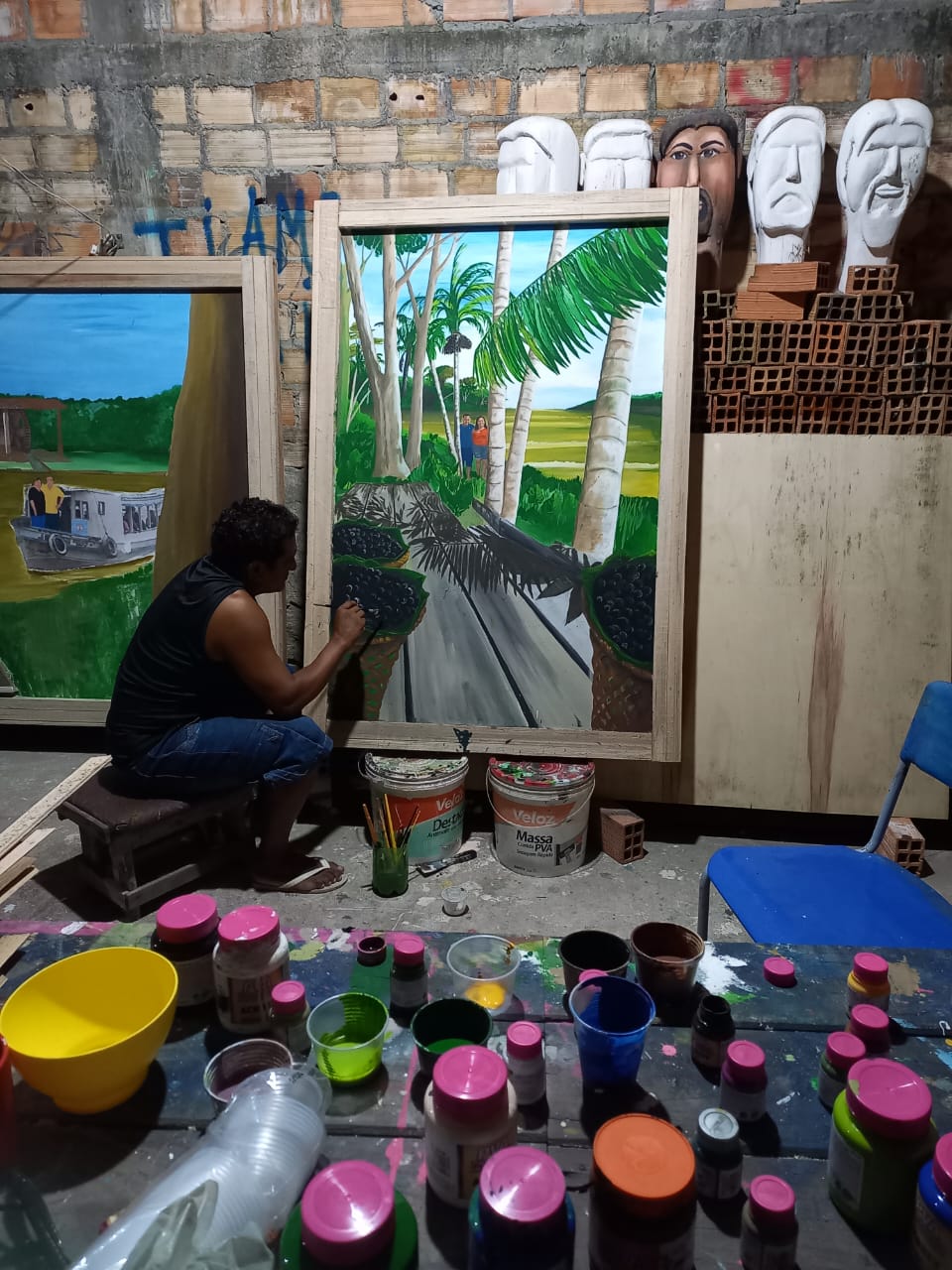 Miguel Barbosa Soares sentado no ateliê, pintando palmeiras e paneiros de açaí. Máscaras de gesso estão na parede de tijolo, e a mesa está cheia de tintas.