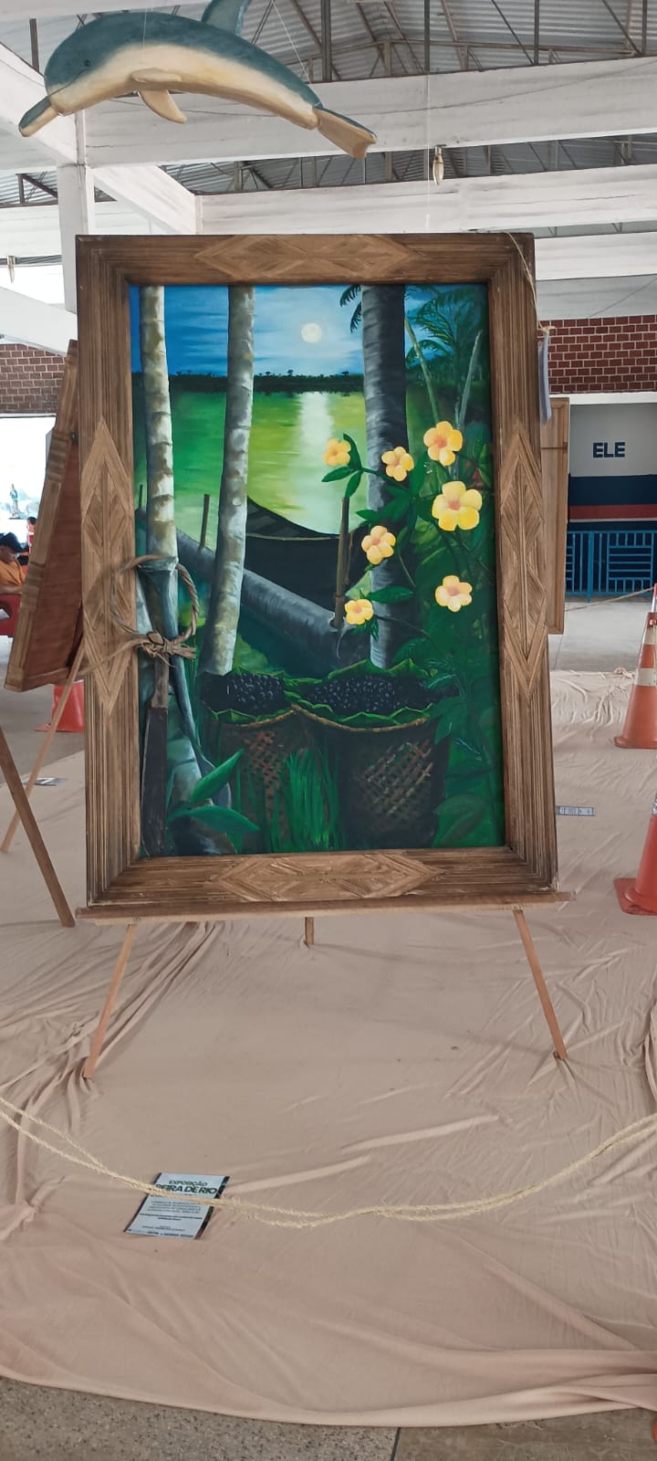 O quadro da canoa, do açaí e das flores amarelas exposto num salão, sobre um cavalete.