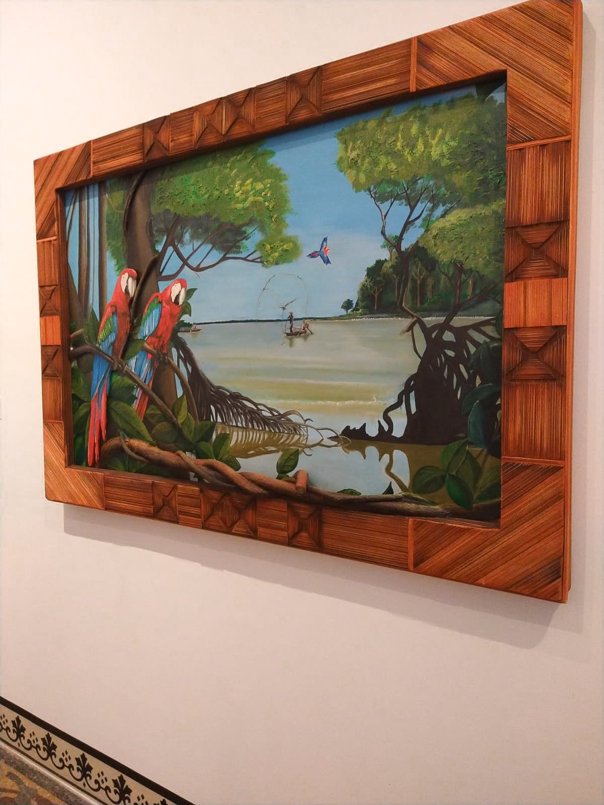 O quadro das araras vermelhas instalado numa parede clara, com a moldura de miriti em relevo.
