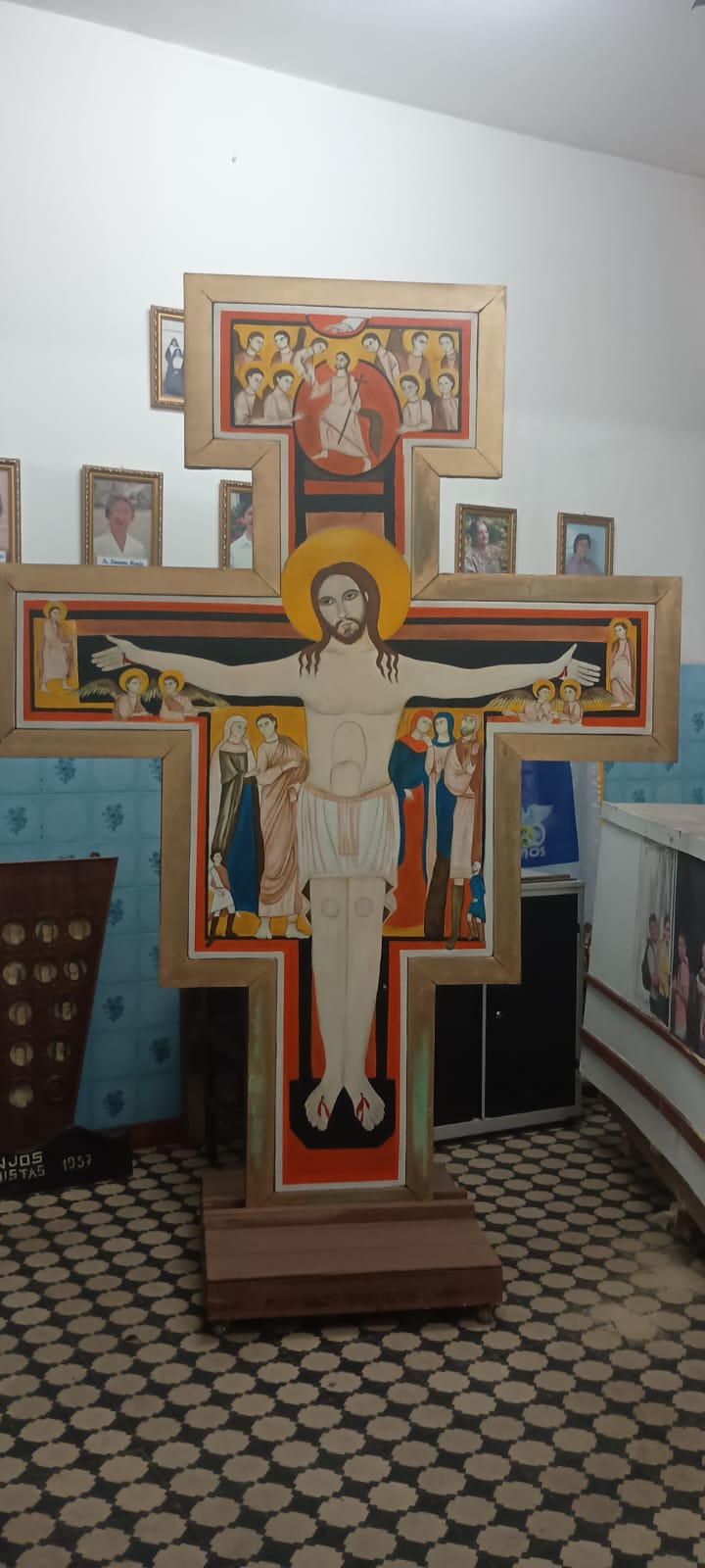 Grande cruz pintada com Cristo de braços abertos, exposta numa sala de piso geométrico.