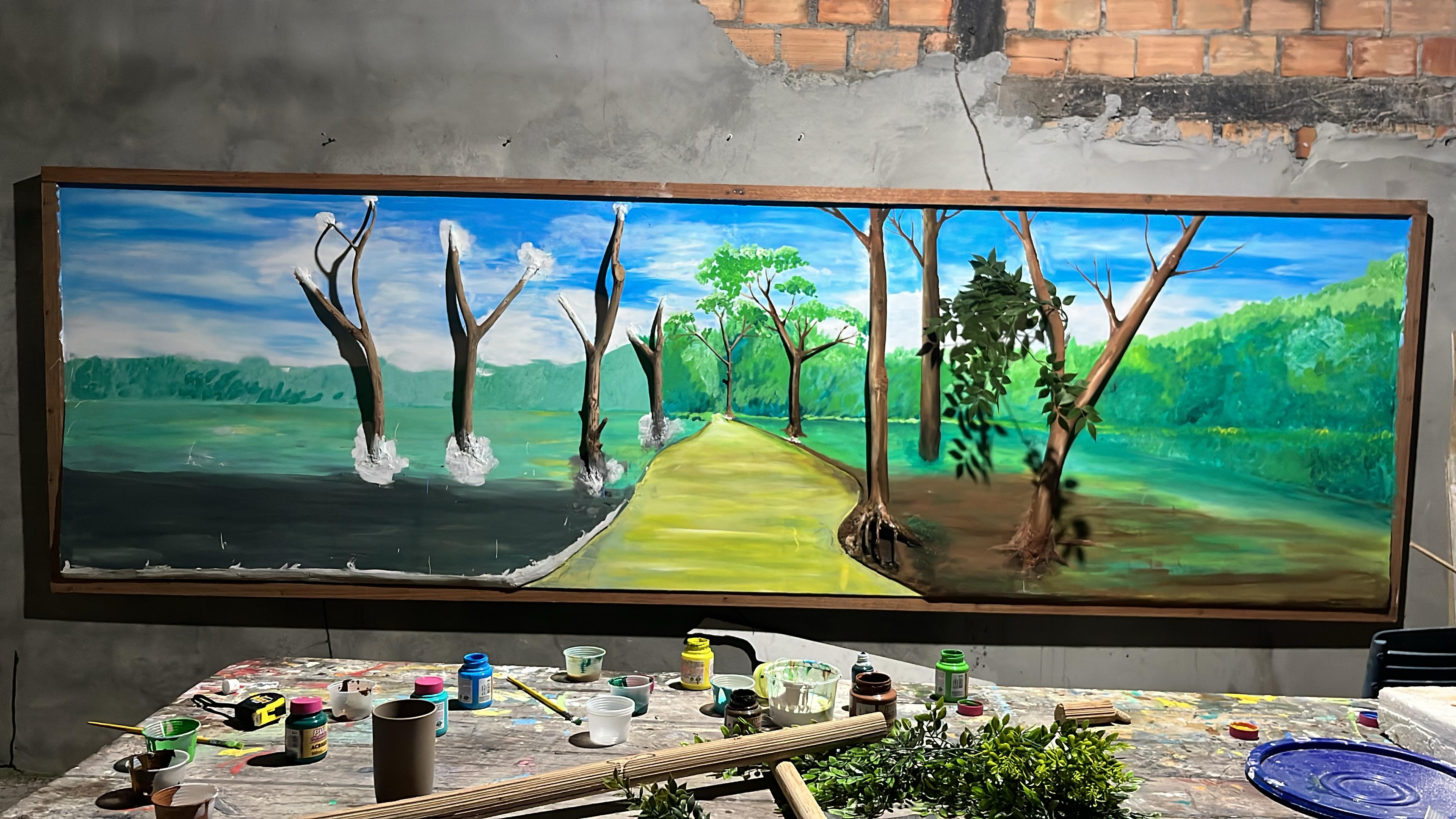 Tela larga de uma paisagem com árvores e um caminho, ainda em pintura, sobre uma mesa coberta de tintas, pincéis e talas de miriti.